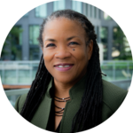 Photo of a black woman in a green blazer. She is standing outside with buildings in the background.