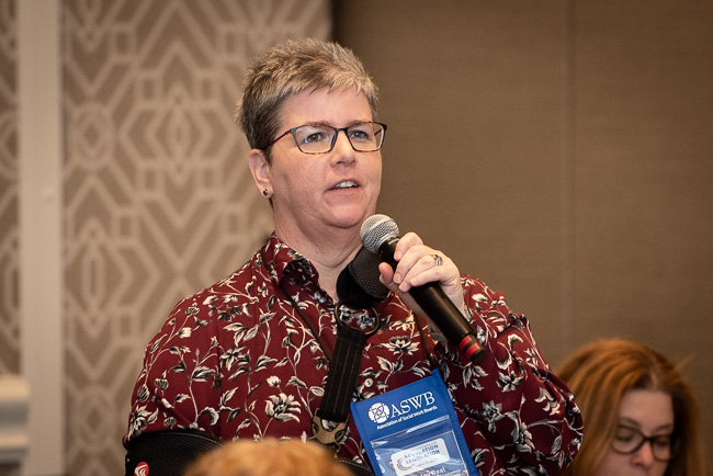 A white woman with short hair and glasses is speaking into a microphone.