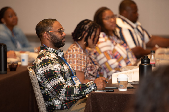 Photograph of four people sitting at a conference table. They are facing a speaker who is outside the frame. One of them is taking notes.