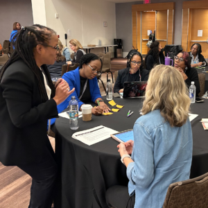 Photograph of a group of women at a table during a conference. There are other tables in the background with groups sitting and talking. Stacey Hardy-Chandler is speaking to the participants in the foreground.