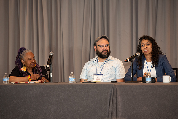 Photograph of Velva Taylor Spriggs, Alejandro Zamora, and Denitha Breau during their panel presentation at the ASWB Education Meeting. They are sitting at a long table with microphones in front of them. Breau, on the far right, is speaking, while Zamora, in the center, and Spriggs, on the left, are listening intently.