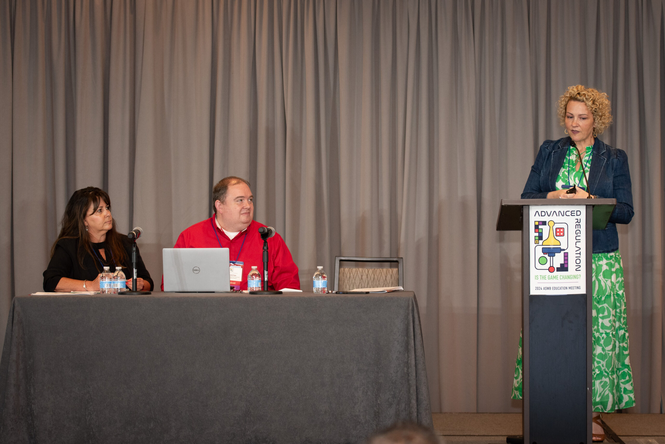 Photograph of panel at a conference. A woman and a man are sitting at a long table on a stage while another woman is standing at a podium speaking.