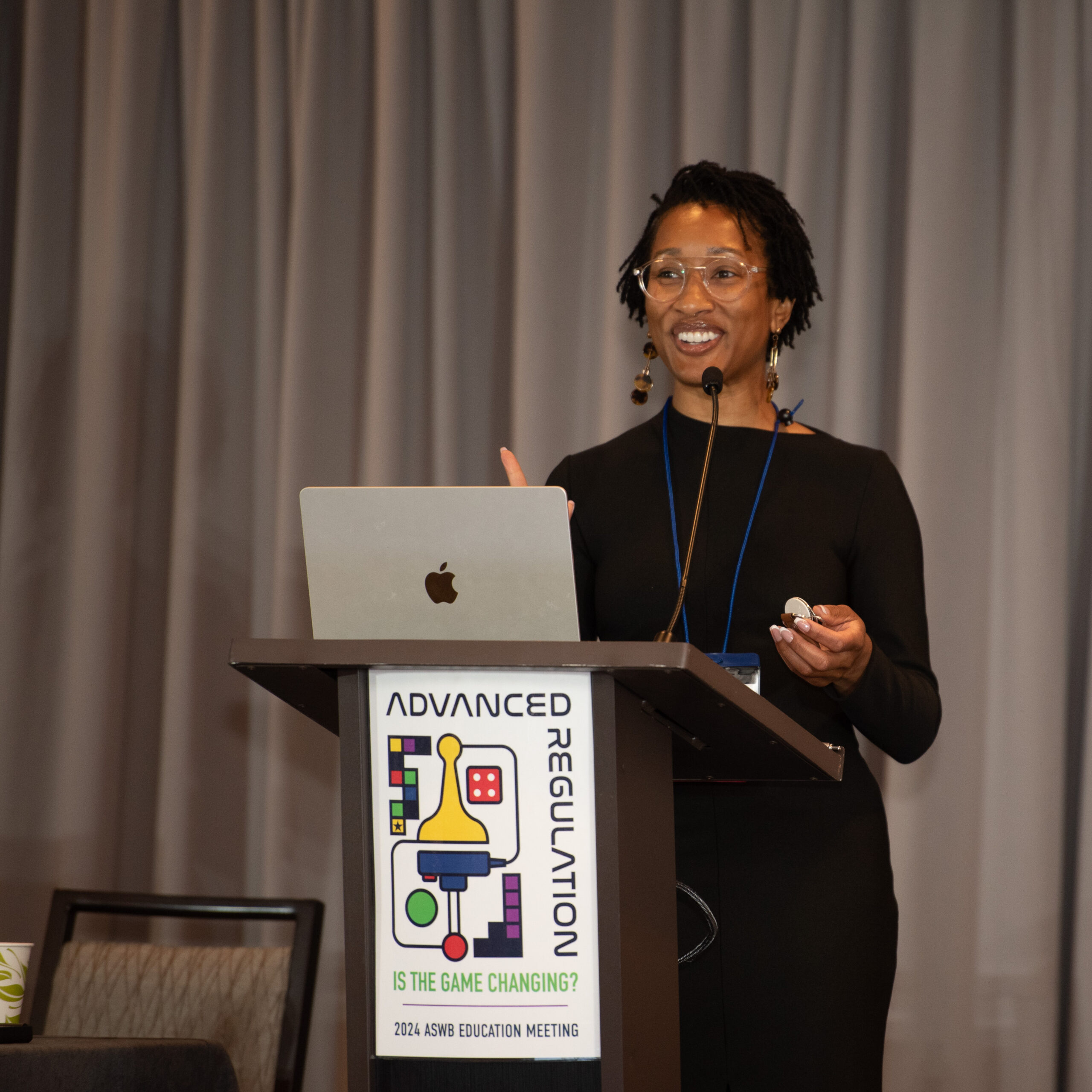 Photograph of a young woman standing at a podium speaking. She is smiling and holding a remote in her hand.
