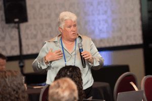 Photograph of Barb Whitenect facilitating a discussion. She is standing with a hand-held microphone and addressing individuals seated in the foreground.