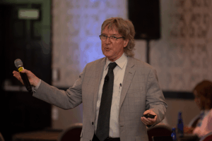 Photograph of Dale Atkinson leading a discussion. He's holding out a hand-held microphone and gesturing to something outside the frame.