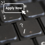 A close-up photograph of a keyboard focused on the question mark button. In the top left-hand corner, the words "Apply Now" are written.