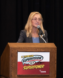 Woman standing behind a podium speaking into a microphone. A sign on the front of the podium reads "Social Work Supervision is a Superpower!"