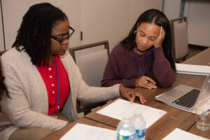 Two women sitting at a desk in a meeting room. They are looking at a packet of papers.