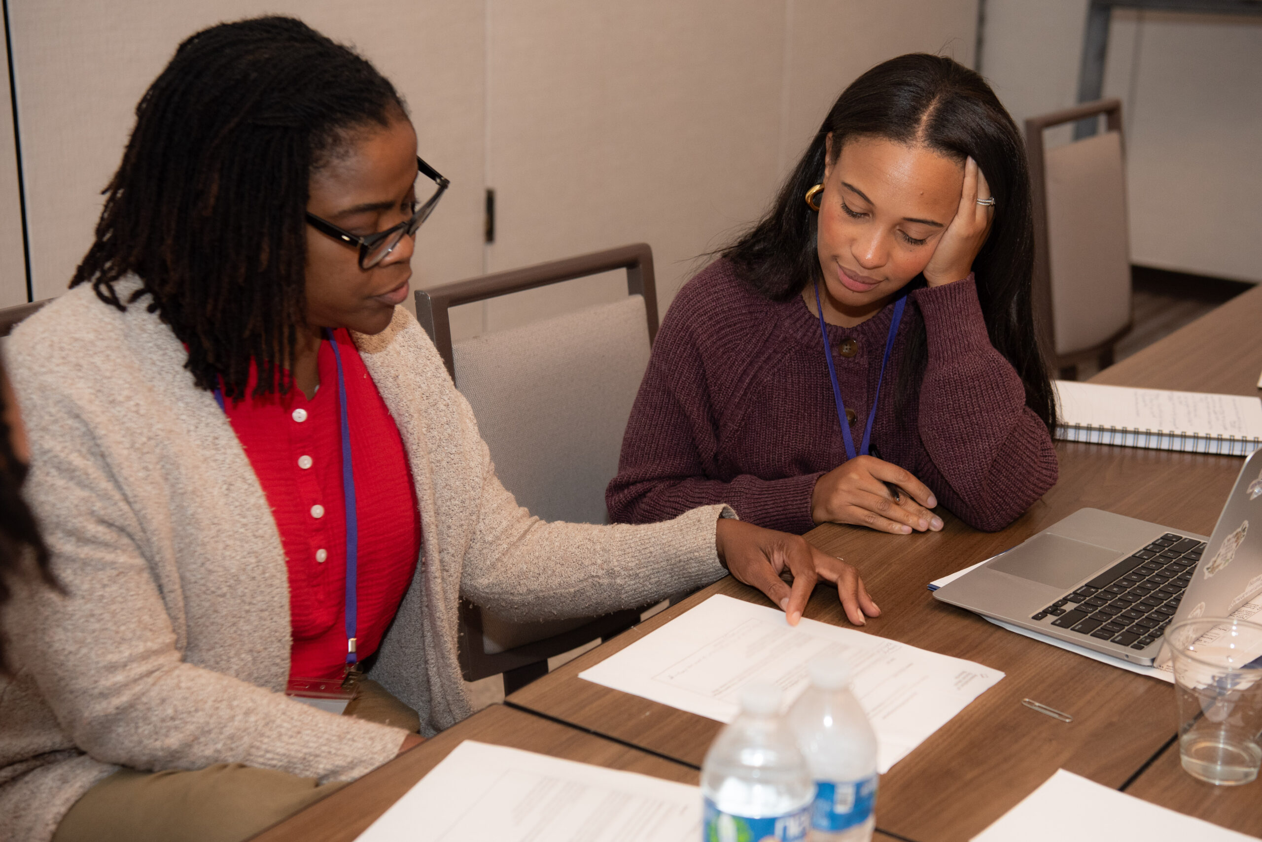 Two women sitting at a desk in a meeting room. They are looking at a packet of papers.